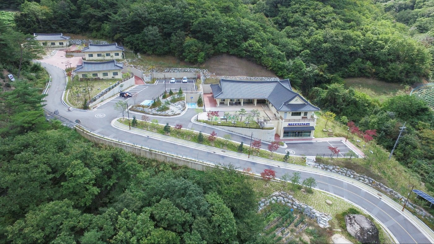 숲으로 둘러싸인 제천한방자연치유센터 전경. 한옥 지붕의 센터동과 숙박동, 굽이진 진입로가 보인다.